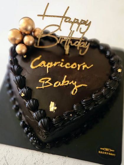 A Belgian Mocha heart-shaped cake for a Capricorn baby. The dark chocolate cake is elegantly decorated with black piping, gold lettering, and a few gold spheres.