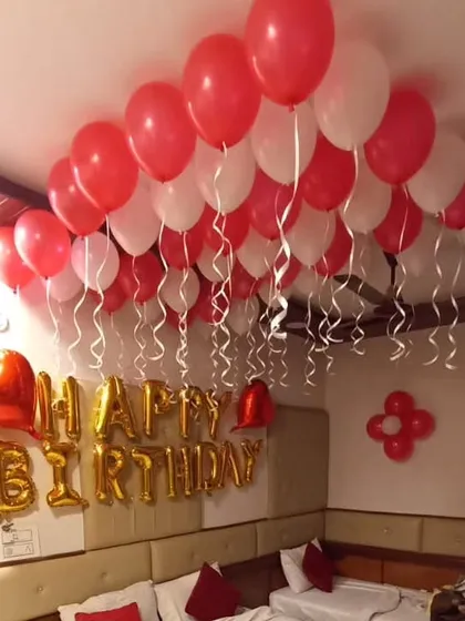 A classic hotel room birthday decoration with red and white ceiling balloons and a gold 'Happy Birthday' banner.
