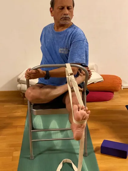A seated hip-opening pose on a chair, using a strap to bind the foot. This is a preparation for Padmasana (Lotus Pose) and other cross-legged postures.