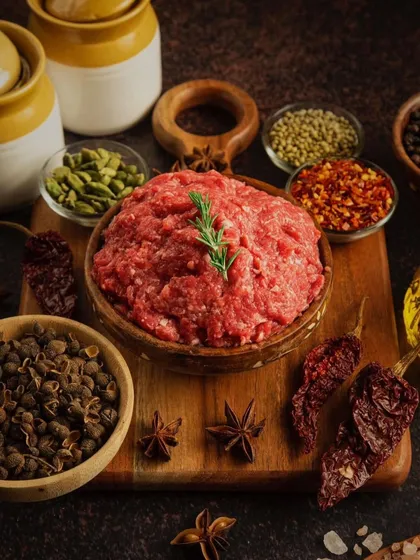 A bowl of fresh minced meat surrounded by a variety of whole spices on a wooden board. The styling evokes a sense of traditional cooking and authentic, bold flavors.