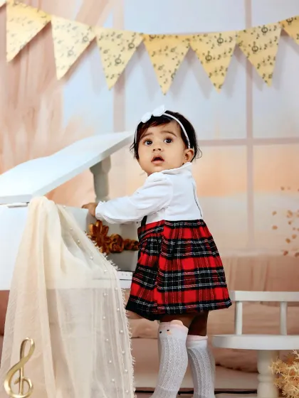A baby girl in a plaid dress stands by a miniature piano, looking up with a curious and sweet expression.