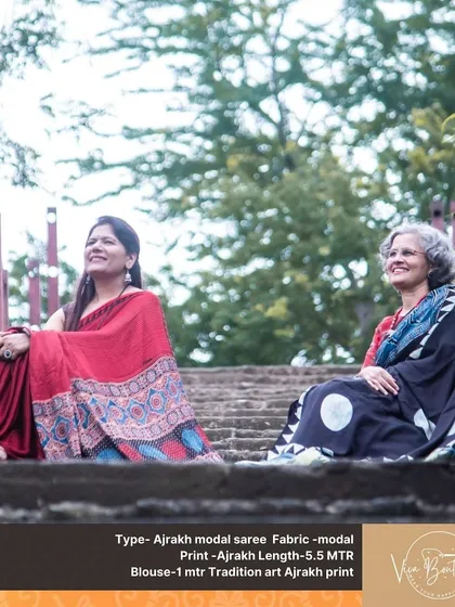 Two happy customers sitting together, both wearing our elegant Ajrakh modal sarees. One is in a classic red, and the other in a striking black with large white motifs.