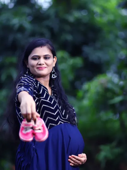 A classic outdoor maternity portrait where the mother-to-be holds up a tiny pair of pink baby shoes, a simple and sweet way to announce the coming arrival of her daughter.