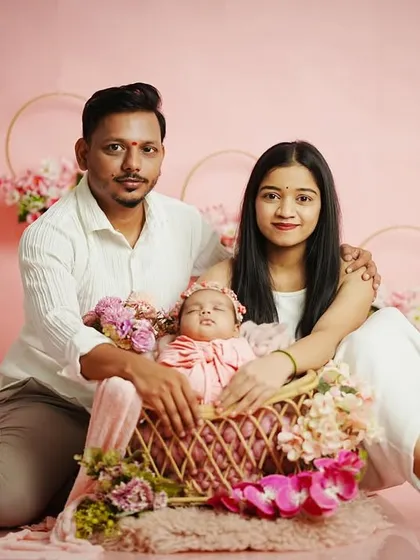 A beautiful family portrait against a soft pink backdrop. The parents cradle their newborn daughter in a flower-adorned basket, creating a sweet and coordinated look. We provide all outfits and props for a stress-free session.
