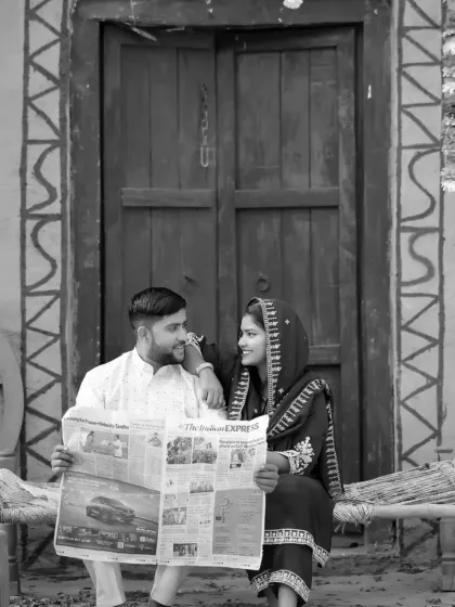 A rustic, village-themed pre-wedding shoot in black and white. The couple is sharing a quiet moment reading a newspaper, creating a timeless and story-driven image.