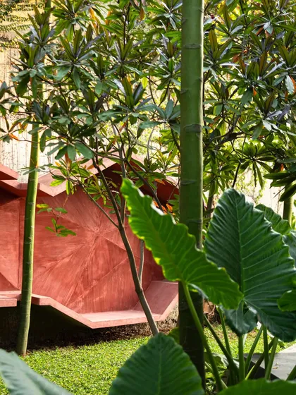 A view of the red sculptural bench through lush foliage. The leaves frame the shot, emphasizing how the man-made art piece coexists with the natural environment.