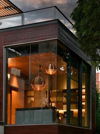 A dusk view of the T House study from the outside. The glass box becomes a lantern in the trees, showcasing the warm interior and the sculptural copper lights, highlighting its role as a special, elevated space.