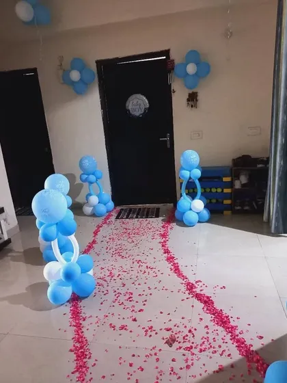 An entrance path for a 'Welcome Baby' party, with blue and white balloon pillars and a walkway sprinkled with red rose petals.