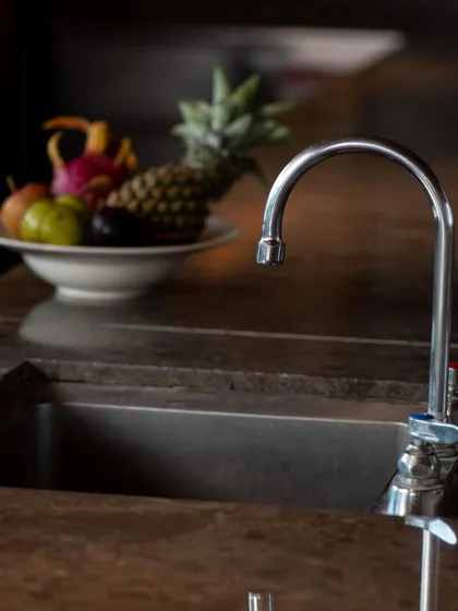 A close-up of a T&S Brass faucet, installed on a dark countertop. The shot highlights the product's chrome finish and functional design in a real-world setting.