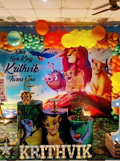 A majestic Lion King theme for a first birthday celebration. This setup features a large backdrop of Pride Rock, character-printed plinths, a 'KRITHVIK' marquee, and a colorful balloon arch to complete the scene.