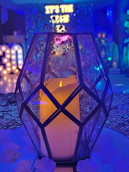 A geometric candle holder centerpiece filled with dried flowers. In the background, the "30" marquee lights and the sequin backdrop are visible, showing how the theme is carried through the space.