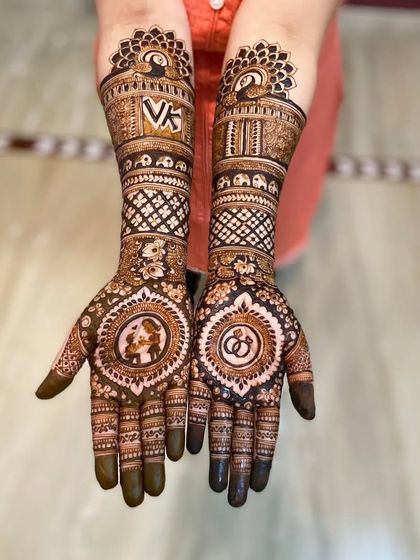 A full-arm engagement design featuring the couple's initials 'VK', a portrait, and interlocking rings. The pattern extends up the arm with traditional motifs like peacocks and elephants.