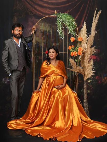A regal and artistic maternity portrait. The mom-to-be is seated in a golden-orange satin gown, framed by a decorative birdcage prop and pampas grass for a unique, painterly effect.