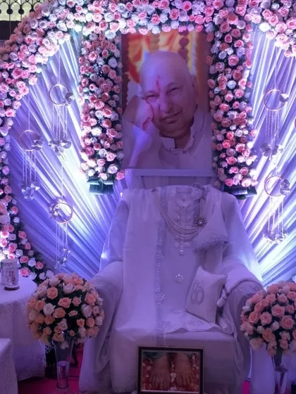A beautiful darbar featuring a circular arch of pink and white roses around Guru Ji's portrait. The gaddi is dressed in pure white, with hanging crystals that catch the light, adding a touch of sparkle to the divine setting.