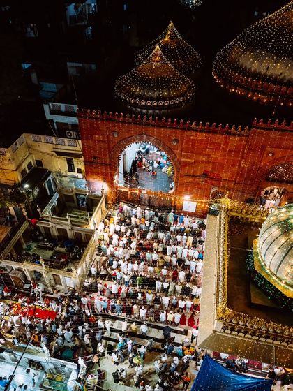 An aerial view of the entrance to Hazrat Nizamuddin Dargah at night, showing crowds gathering for prayers. The illuminated domes and the warm glow from the entrance create a welcoming and spiritual ambiance.