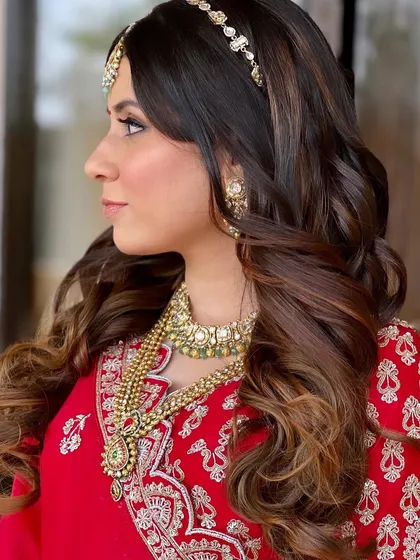 A side profile of this beautiful bridal look, showcasing the soft curls and the elegant matha patti. The hairstyle is designed to complement the traditional red saree.