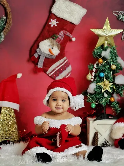 A little Santa's helper getting ready for Christmas. The festive red background, tree, and Santa Claus doll make this a perfect holiday-themed shoot.