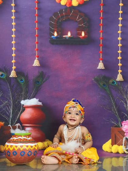A baby dressed as Little Krishna sits happily in a festive Janmashtami setup. The marigold garlands, peacock feathers, and clay pots create an authentic and vibrant scene.