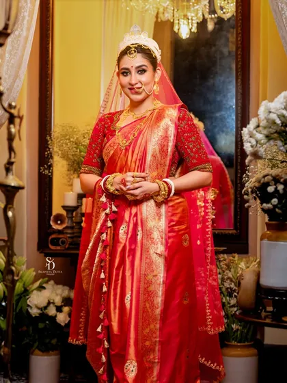Actress Oindrila Sen dressed as a traditional Bengali bride. The warm lighting and opulent setting create a classic portrait, highlighting the red Banarasi saree and the customary 'sholar mukut'.