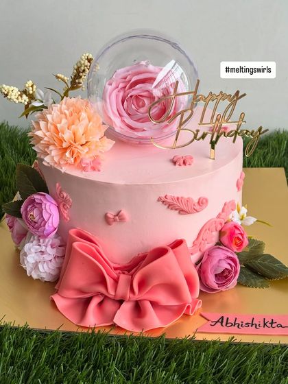 A dreamy pink whipped cream cake perfect for a teen's birthday. It's decorated with a large handmade fondant bow, artificial flowers, and a delicate rose inside a clear sphere.
