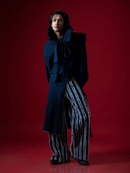 A full-length portrait of a male model in an avant-garde, high-fashion outfit against a bold red background.