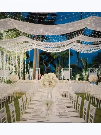 An all-white themed reception that exudes pure elegance. The clean lines of the tables, chairs, and cascading floral arrangements under a ceiling of fairy lights create a dreamy, romantic setting.