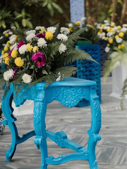 A brightly painted blue console table holds a vibrant floral arrangement, adding a pop of color and vintage charm to the Haldi decor.