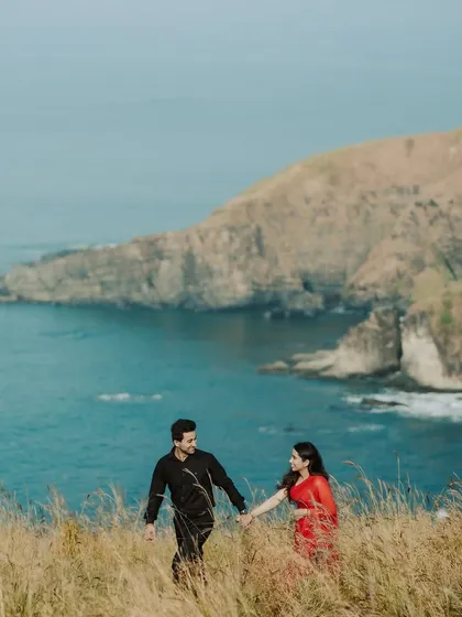 A couple walks hand-in-hand through tall grass on a cliff overlooking the sea. The vibrant colors of their outfits against the natural landscape create a stunning, romantic image.