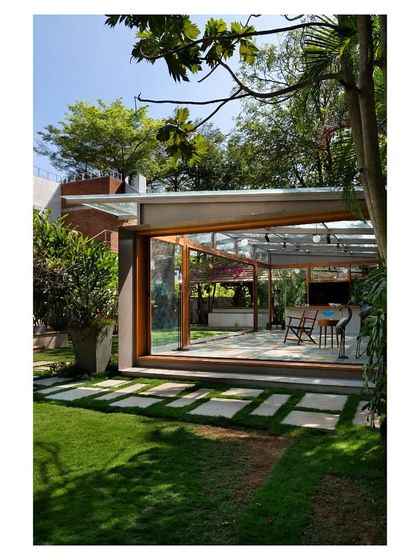The Komorebi Pavilion seen from the garden, appearing as a light-filled lantern in the landscape. The design uses a minimal steel frame and extensive glazing to dissolve the building's mass, allowing it to merge with the surrounding trees and pathways.