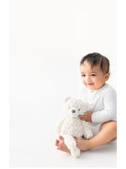 A happy six month old cuddling his teddy bear. The simplicity of the white background and ribbed onesie keeps the focus on his sweet smile and the comforting bond with his fluffy friend.