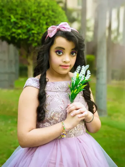 A young girl dressed in a beautiful lavender gown holds a sprig of flowers, looking like a princess in a magical garden.