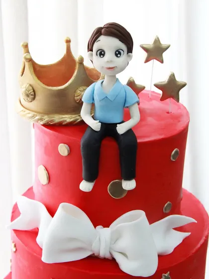 A close-up of the handcrafted boy topper and golden crown on the vibrant red birthday cake.
