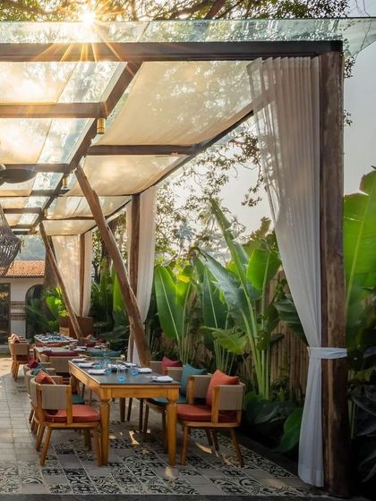 A sun-drenched dining patio at Sanctuary Bar, with tables set under a glass and fabric canopy. The design allows for comfortable dining while still feeling completely immersed in the lush garden setting.