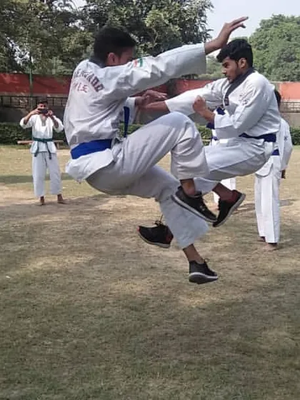 Two students practice a jumping kick during a sparring session in a park. Outdoor training adds a new dimension to their practice.
