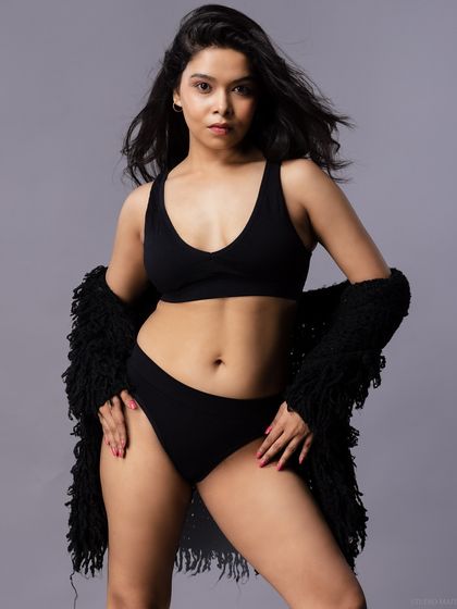 A confident pose in a simple black two-piece lingerie set, paired with a textured black shrug. The grey background keeps the focus entirely on the model.