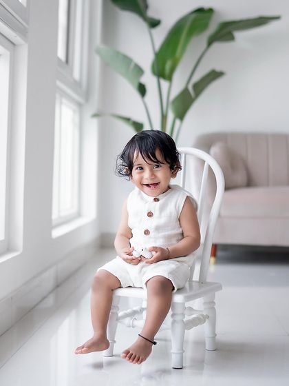 A smiling baby boy sitting on a small white chair in the studio.