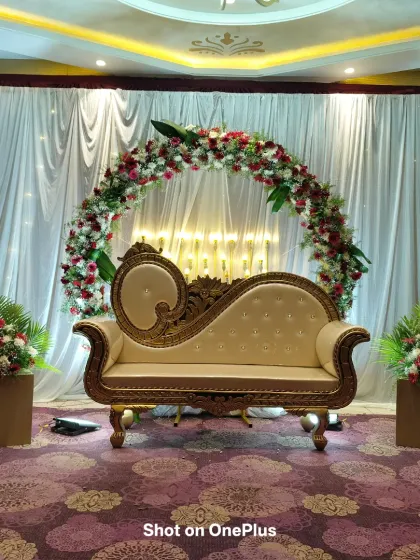 A classic half-saree function setup with a red and white floral arch against a shimmery white curtain. The golden-framed diwan provides a royal seating arrangement for the ceremony.