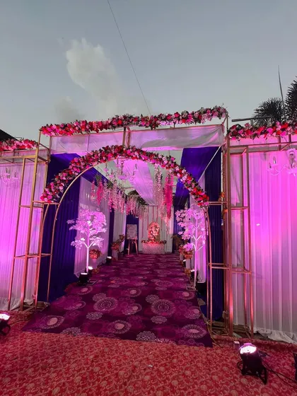 An outdoor wedding entrance with a beautiful floral arch. The purple and white drapes combined with pink lighting create a colorful and celebratory welcome.