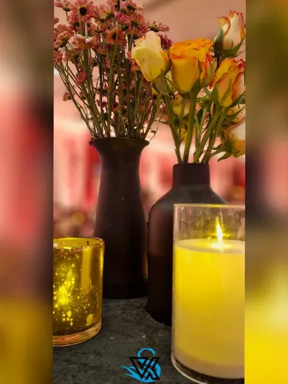 A close-up of a table centerpiece, featuring yellow roses and dried flowers in black vases, with the warm glow of a candle adding to the moody, autumnal ambiance of the cocktail party.
