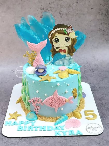 A cute mermaid cake featuring a mermaid character, a pink fish, and a translucent blue sail to mimic water.