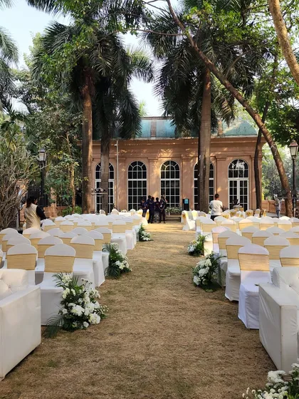 An elegant outdoor ceremony setup with white sofas and chairs arranged on a lush lawn, facing the beautiful venue facade.