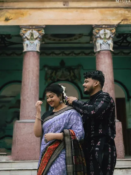 A tender moment of affection. He gently places a flower in her hair as she smiles, standing before the weathered, ornate pillars of a heritage building in Sambalpur.