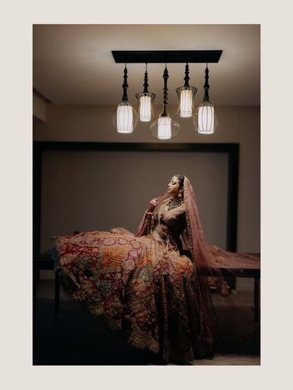 A regal portrait of Radhika seated, with her elaborate lehenga spread out. The dramatic lighting from above adds a touch of old-world glamour and sophistication.