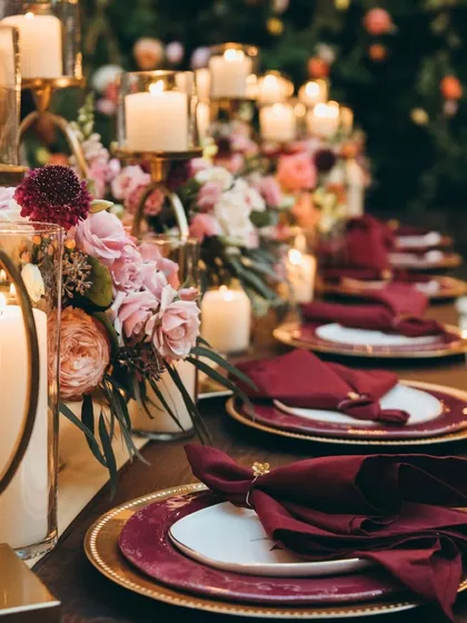An opulent and romantic dinner setting. The table is aglow with numerous candles in hurricane glasses and candelabras, surrounded by lush arrangements of burgundy and pink flowers.