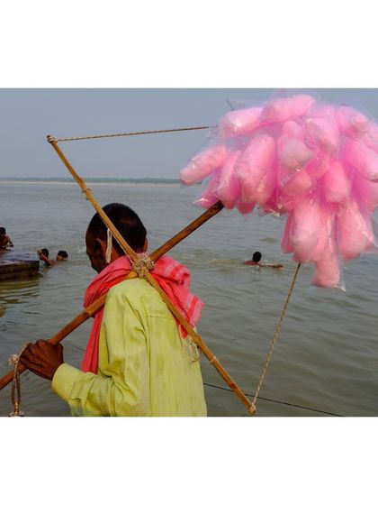 A candy floss seller walks along the banks of the Ganga in Varanasi. The bright pink of the candy contrasts with the earthy tones of the river and the people bathing in the background.
