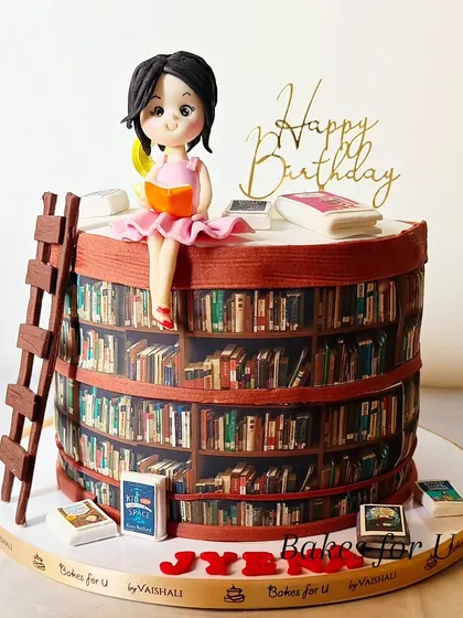 A book lover's dream cake, designed to look like a circular library. The cake is wrapped in an edible image of bookshelves and topped with a fondant figure of a girl reading.