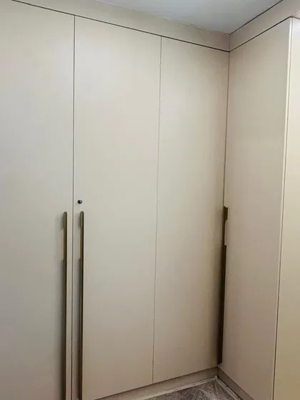This L-shaped wardrobe features a premium matte laminate finish in a neutral beige tone. The custom-made long external handles in a brass finish add a touch of elegance.