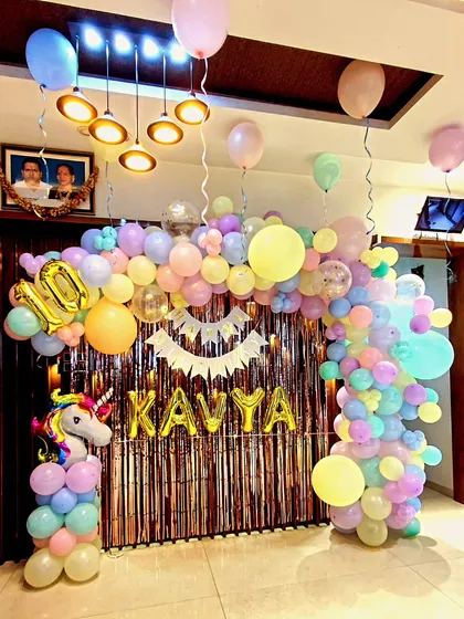 A dreamy unicorn party decoration for a tenth birthday, using a soft pastel color palette. This design features an organic balloon garland in pastel pink, yellow, blue, and mint, a shimmering backdrop, and a large unicorn figure. The birthday girl's name "KAVYA" is spelled out in gold foil balloons, adding a personalized touch to this magical setup.