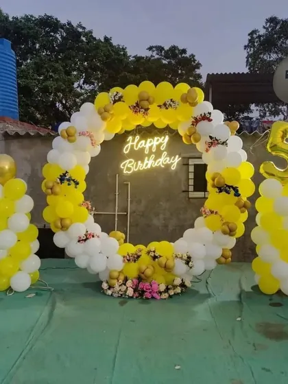 A birthday party on a terrace with a circular balloon arch in yellow and white, and a 'Happy Birthday' neon sign.