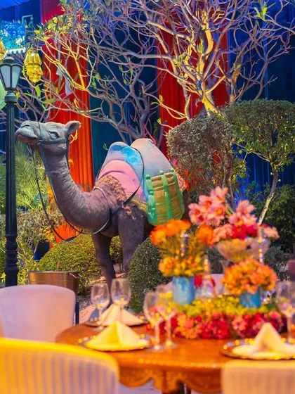 A vibrant tablescape from an Aladdin-themed birthday party. The scene includes a life-sized camel prop, colorful floral arrangements, and Moroccan-style lanterns, contributing to the immersive desert oasis feel.